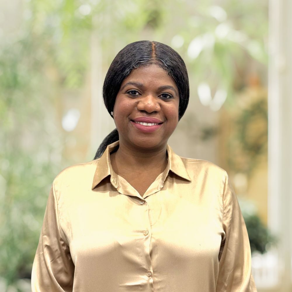 A stylish woman wearing a gold shirt, standing gracefully beside a plant.