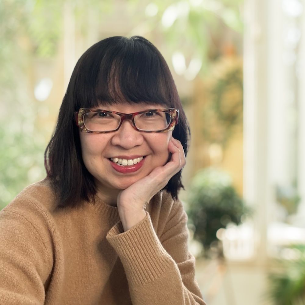 A woman with glasses and a brown sweater smiling, radiating warmth and friendliness.