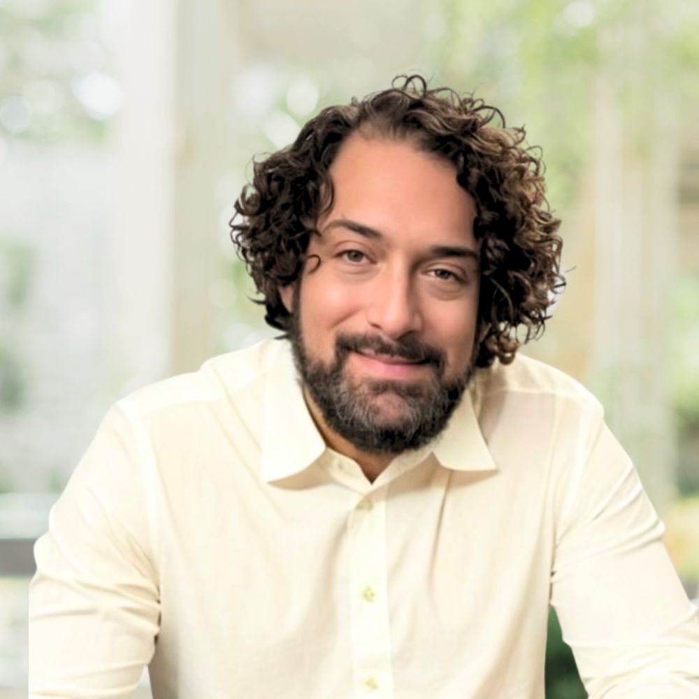 A man with curly hair and a beard wearing a white buttoned up shirt.