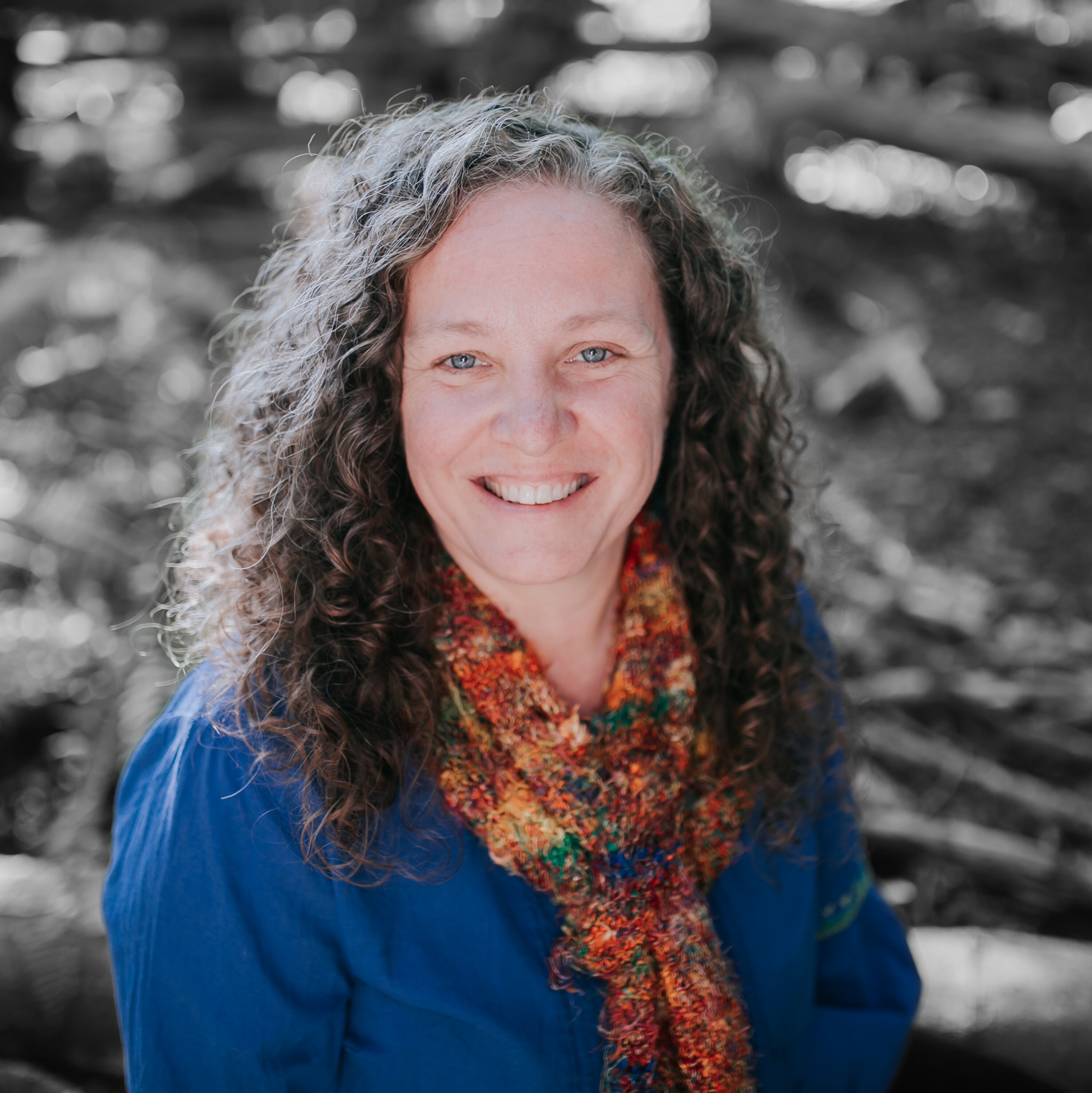 A professional headshot of a counselor woman wearing a blue outfit and a vibrant, multi-colored scarf. The background features a blurry forest, adding a natural and calming atmosphere to the image.